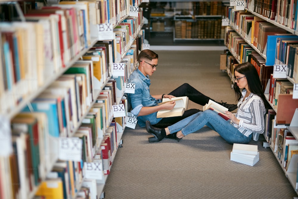 Estudantes que estudam na biblioteca da universidade. Homem e mulher lendo livros sentados no chão entre estantes. Alta Resolução