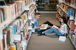 Estudiantes que estudian en la Biblioteca Universitaria. Hombre Y Mujer Leyendo Libros Sentados En El Suelo Entre Estanterías. Alta resolución