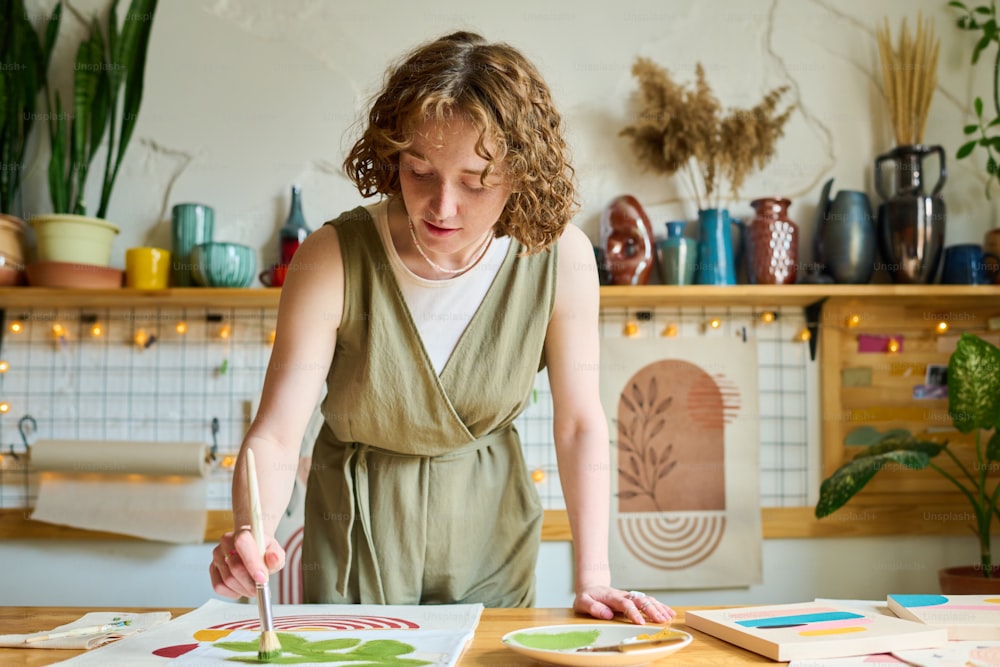 Junge kreative Frau, die sich über den Tisch beugt, während sie abstraktes Bild auf Papier mit Pinsel und grünem Aquarell im Workshop malt