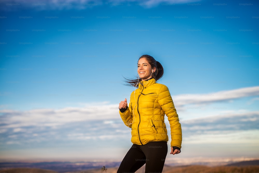 Adorable happy smiling sporty girl jogging outside in winter sportswear with earphones.