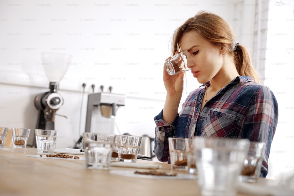 Portrait in selective focus of young female barista examining cups with beans and ground coffee, doing cupping test, smelling fresh coffee