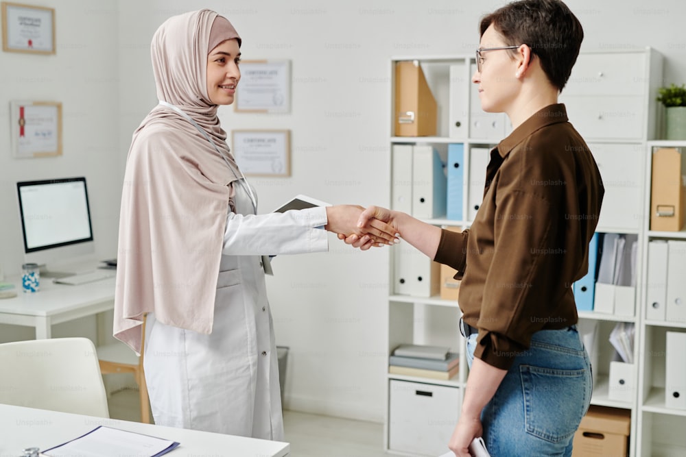 Happy young females shaking hands in medical office by doctor workplace while congratulating one another on good results of treatment