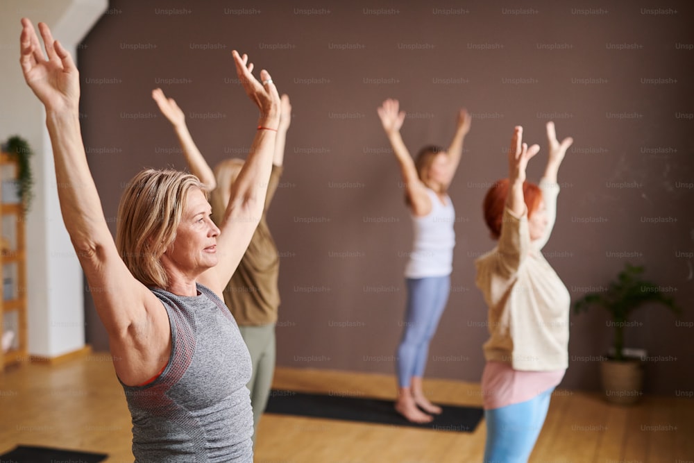 Group of people raising their hands up doing stretching exercises while standing on exercise mat in wellness club
