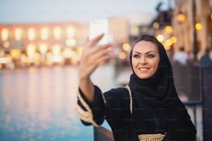 Belle fille avec hijab prenant un selfie souriant à l’extérieur dans la ville près de la rivière.