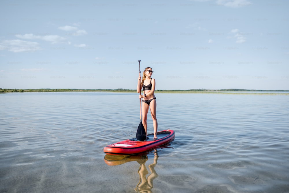 Portrait of a young woman in black swimsuit paddleboarding on the lake with calm water during the sunset