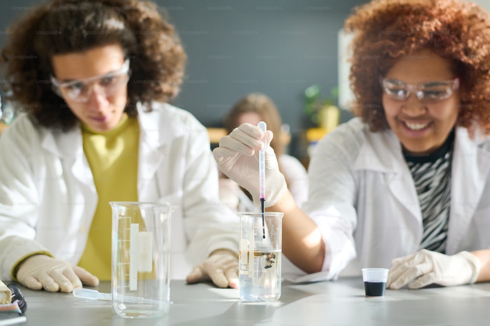 化学の授業で、手袋、眼鏡、白衣を着た10代の女子学生が、透明な液体が入ったチューブに黒い液体物質を滴下する