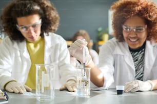 Teenagerin in Handschuhen, Brille und Laborkittel lässt schwarze flüssige Substanz in Röhre mit transparenter Flüssigkeit fallen im Chemieunterricht