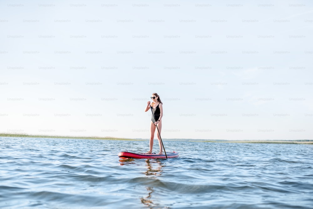 Bella giovane donna in costume da bagno nero paddleboarding sul lago con le onde durante la luce del mattino