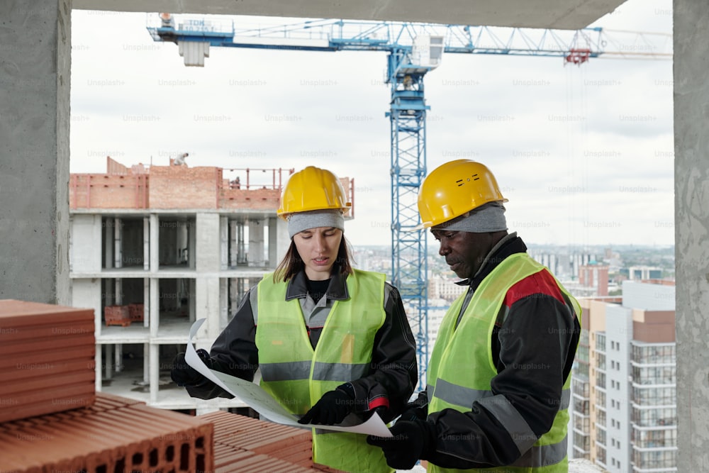 Jeune femme constructeur et contremaître expérimenté portant des casques de sécurité et des vêtements de travail discutant d’un croquis sur un plan par pile de matériaux de construction