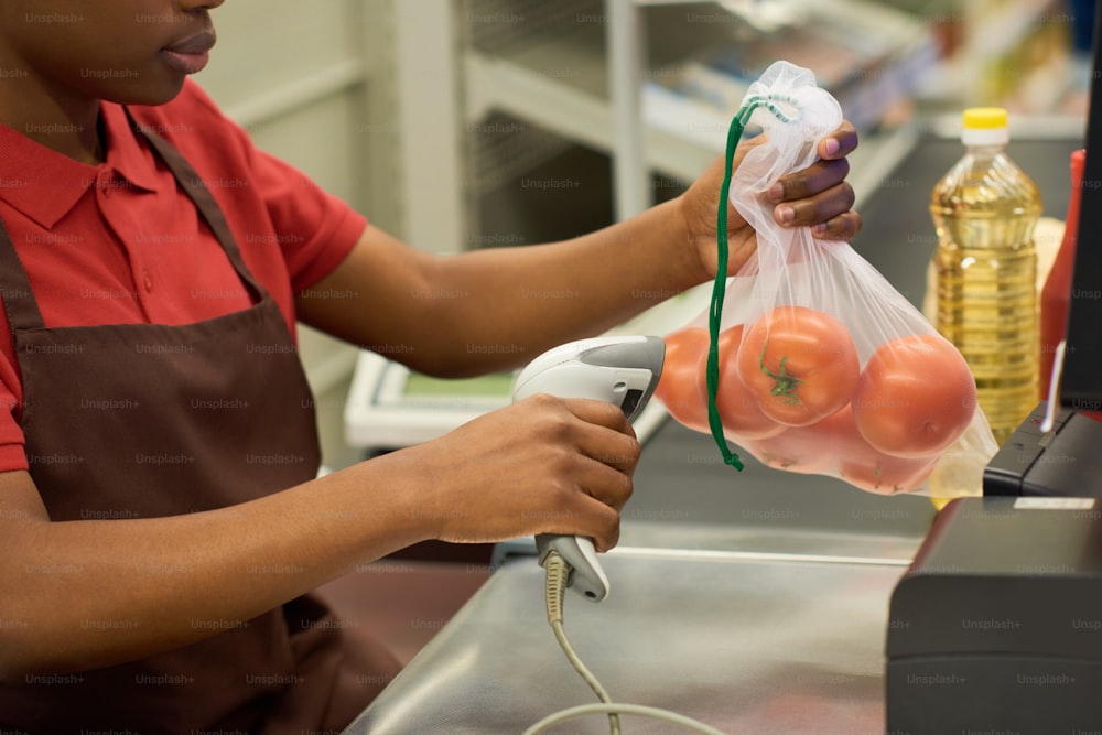 Mujer negra joven con camisa roja y delantal marrón escaneando tomates frescos en bolsa de celofán sobre el mostrador del cajero en el supermercado