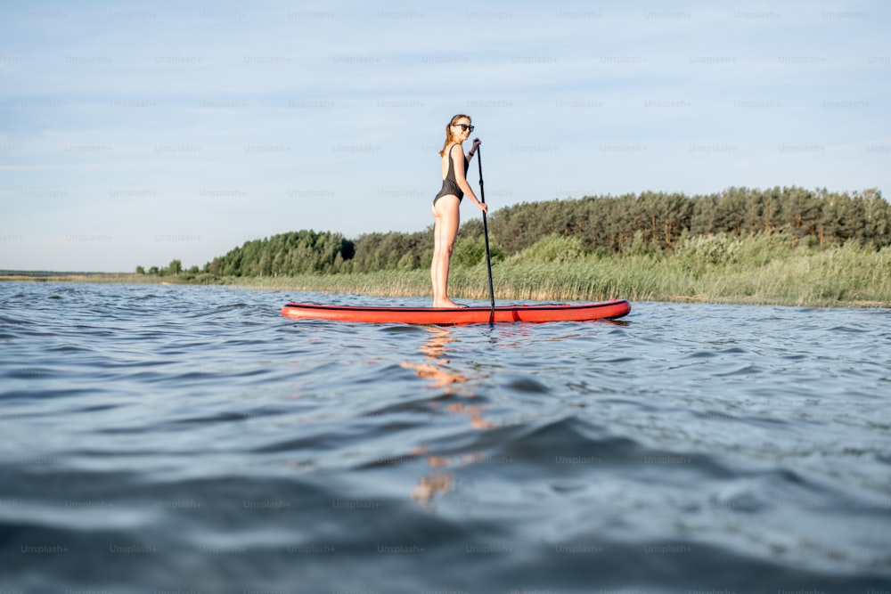 Beautiful young woman in black swimsuit paddleboarding on the lake during the morning light