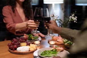 Hands of young interracial dates clinking with glasses of red wine over dinner table served with healthy food and snacks