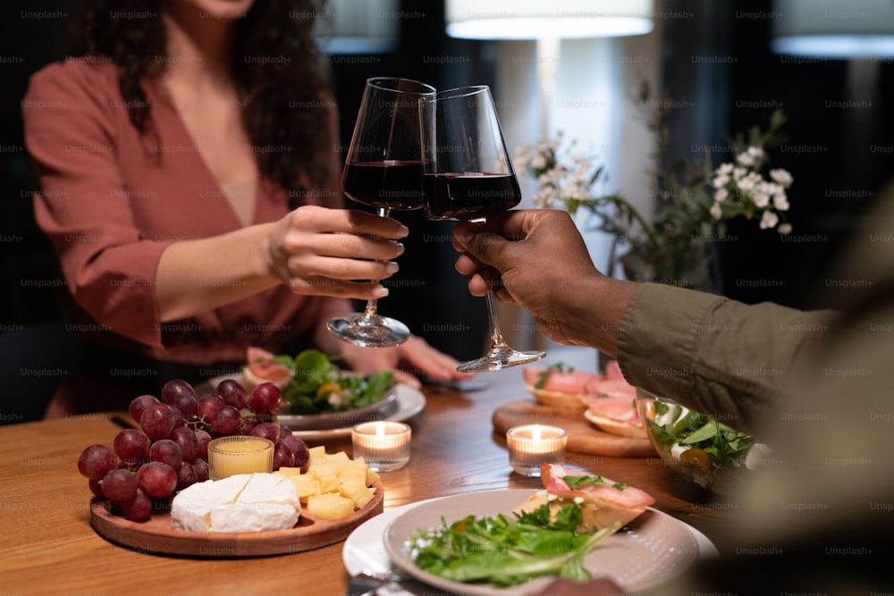 Hands of young interracial dates clinking with glasses of red wine over dinner table served with healthy food and snacks
