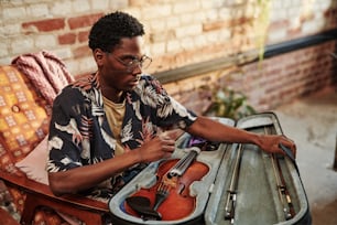 Junger schwarzer Mann in stilvoller Freizeitkleidung, der den Bezug mit Geige und Fiddlesticks öffnet, während er im Sessel im Wohnzimmer der Loft-Wohnung sitzt