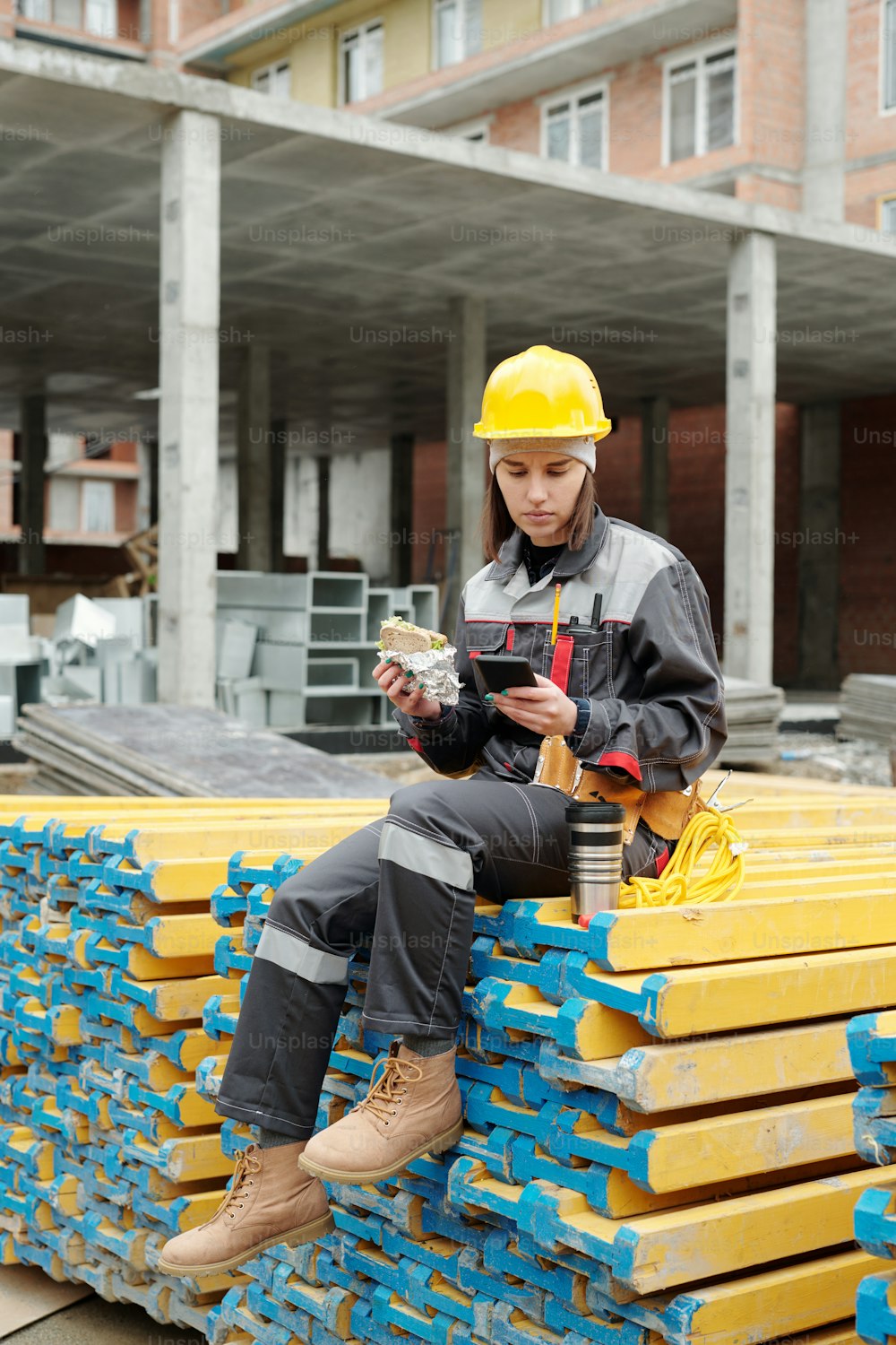 Young female builder in workwear having sandwich and using smartphone while sitting on stack of planks at lunch break