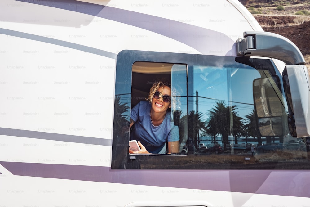 Happy woman portrait smiling at the window of her modern camper van motor home. Concept of tourist and travel for holiday vacation or van life lifestyle. Young adule female people happy at destination