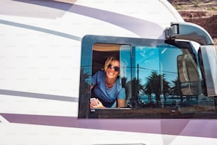 Happy woman portrait smiling at the window of her modern camper van motor home. Concept of tourist and travel for holiday vacation or van life lifestyle. Young adule female people happy at destination
