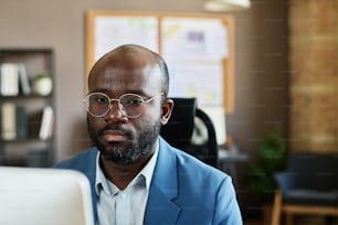 African serious businessman in eyeglasses looking at computer monitor and concentrating on his work at office
