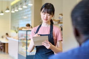 Young Asian waitress in apron making notes in notepad while taking order from man at restaurant