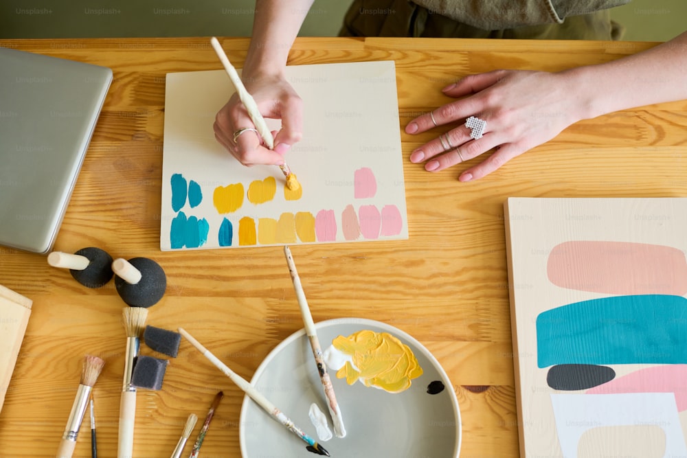 Above view of hands of young creative woman making color swatches on paper while sitting by table and working over abstract painting