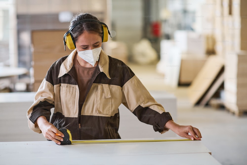마스크와 유니폼을 입은 젊은 여성 노동자가 공장에서 일하는 동안 줄자로 측정을 하고 있다