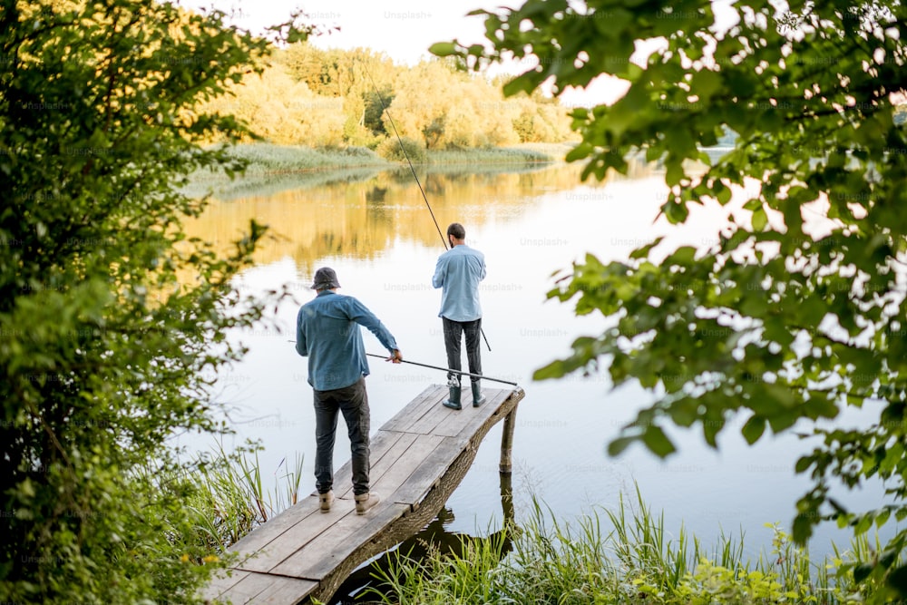 Dos amigos varones pescando juntos de pie en el muelle de madera durante la luz de la mañana en el hermoso lago
