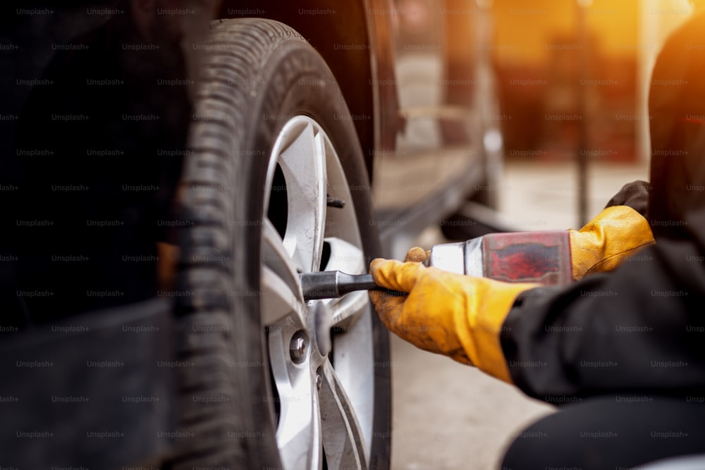 An experienced hard working mechanic in orange gloves is using an electric wrench to tighten up the screws in a car wheel.