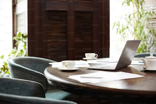 Large wooden table with tea cups, laptop and papers in empty meeting room