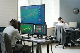 Hombres afroamericanos y caucásicos sentados frente a monitores de computadora trabajando con datos, analizando estadísticas monetarias y haciendo llamadas telefónicas