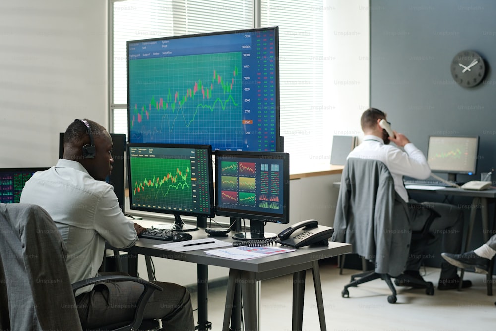 African American and Caucasian men sitting in front of computer monitors working with data, analyzing currency stats and having phone calls