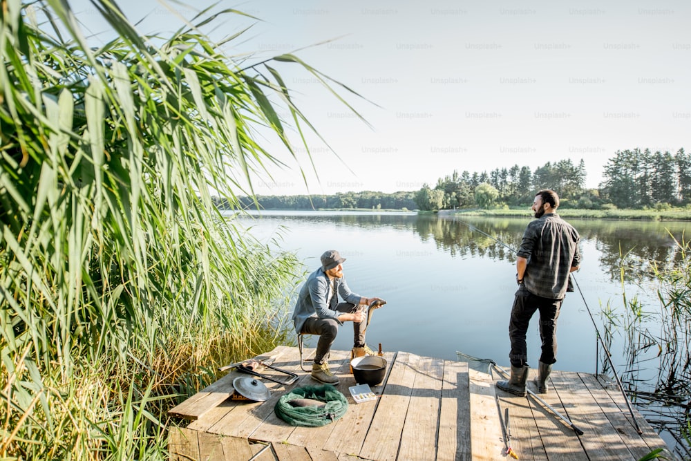 아침에 호수에 녹색 갈대가 있는 아름다운 나무 부두에서 피크닉을 즐기는 동안 휴식을 취하는 두 명의 어부