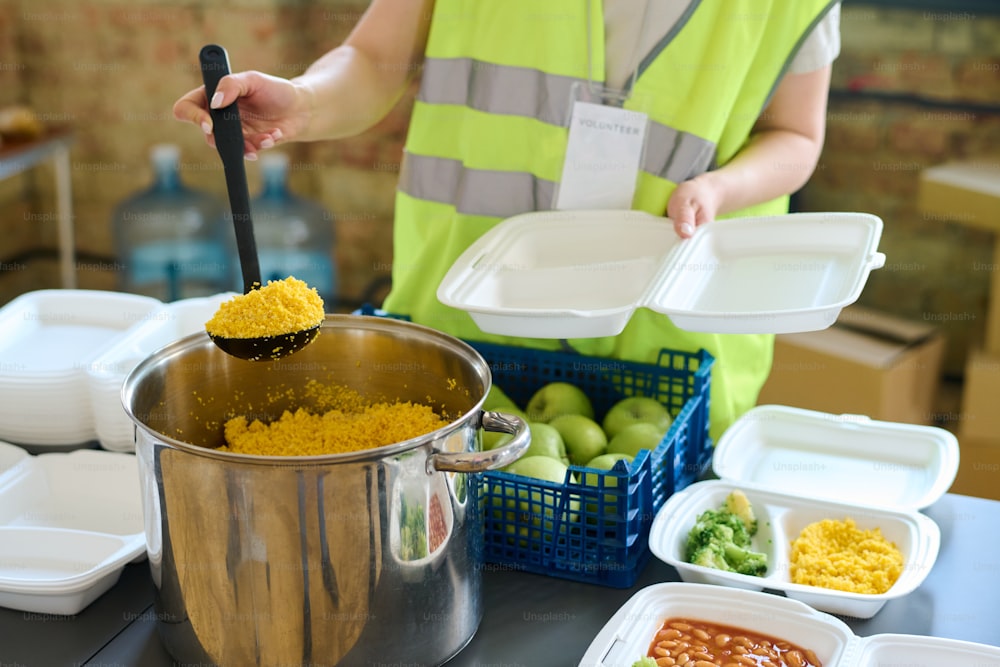 Joven voluntaria en uniforme sosteniendo un cucharón con comida cocinada en una sartén grande antes de ponerla en un recipiente de plástico para los refugiados