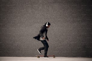 Elegante jovem skatista hipster dreadlock com fones de ouvido brincando com skate perto da parede cinza.