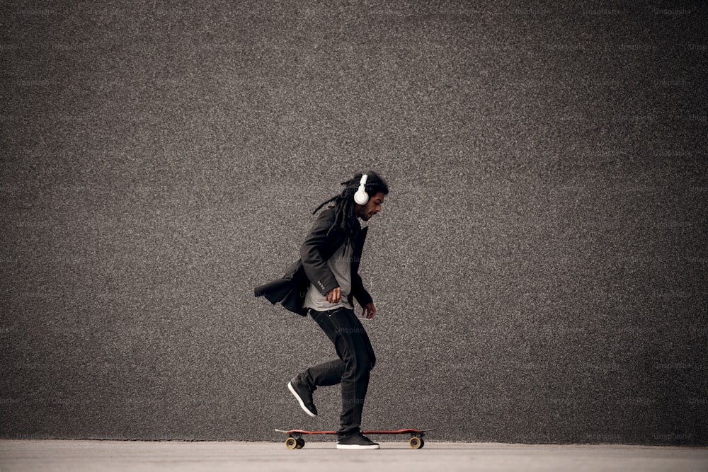 スタイリッシュな若いドレッドヘアのヒップスタースケーターは、灰色の壁の近くでスケートで遊んでヘッドフォンを持っています。