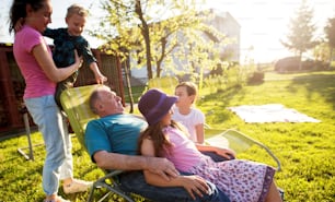 Grandfather enjoys a sunny day on a field near the house while being surrounded by adorable grandchildren while one of the girls is sitting in his lap.
