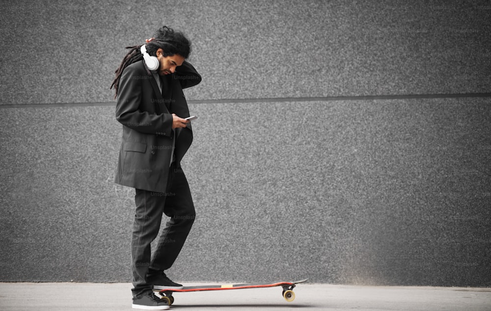 Primer plano de un elegante hombre patinador con rastas feliz y guapo en un traje de pie sobre un patín con una pierna mientras lee un mensaje de un móvil en la calle.