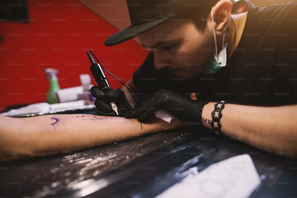 젊은 집중 문신 예술가는 그의 가게에서 고객의 팔을 조심스럽게 잉크로 칠하고 있습니다.