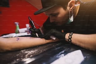 Young focused tattoo artist is inking customers arm carefully in his shop.