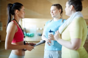 Glückliche übergroße Frau mit einer Flasche Wasser, die zwischen den Trainingseinheiten Fragen an den Fitnesstrainer stellt