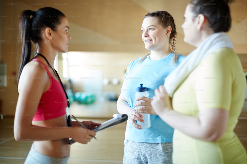 Mujer feliz de gran tamaño con una botella de agua que hace preguntas al instructor de fitness entre entrenamientos