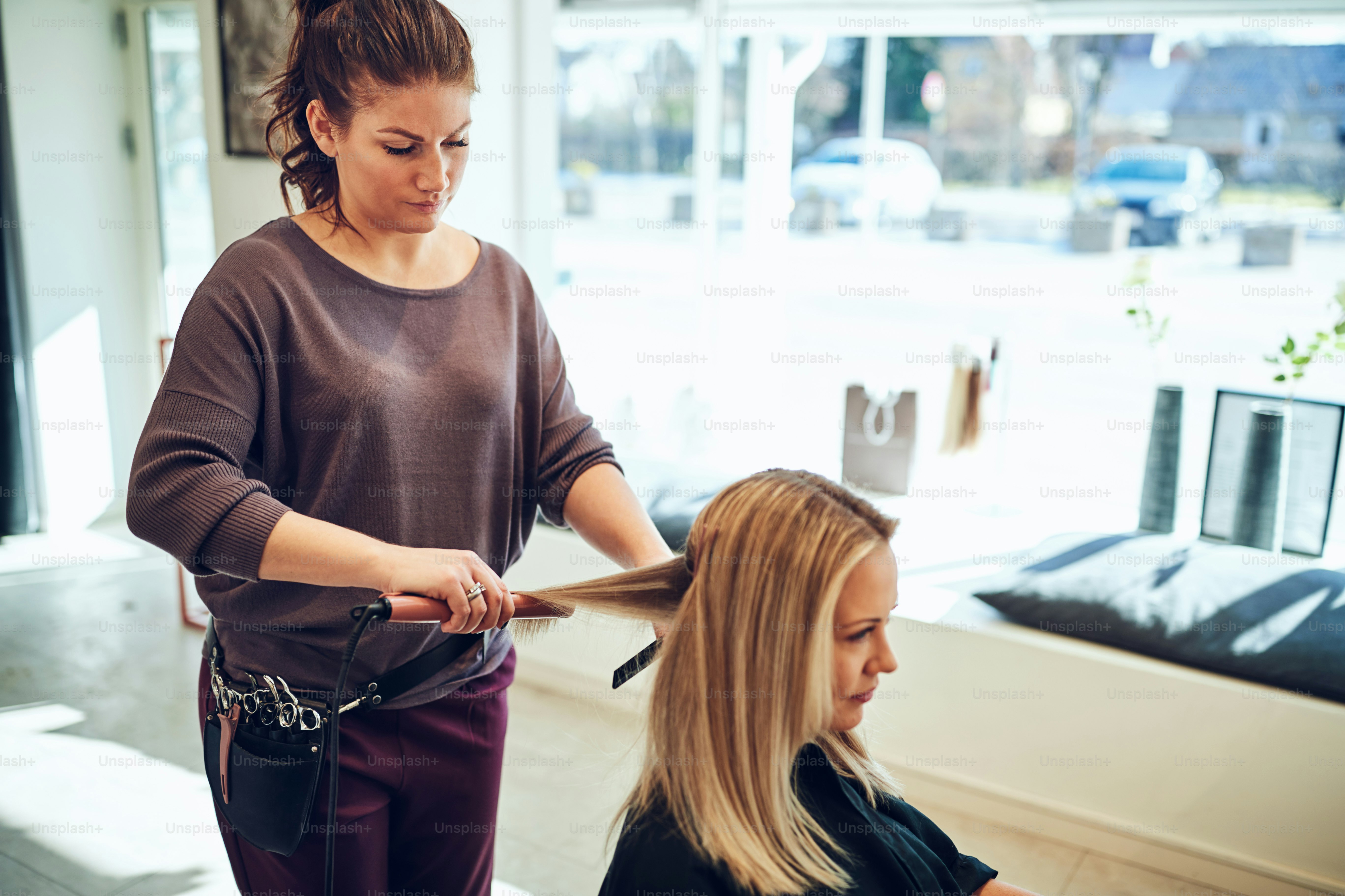 Young blonde woman sitting in a salon chair getting her long blonde hair straightened by her hairstylist