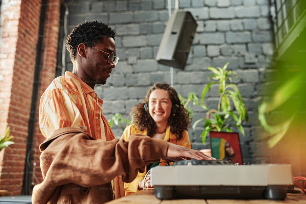 Hombre negro feliz ajustando el controlador de dj mientras crea nueva música contra su alegre novia mientras pasa tiempo con ella en casa