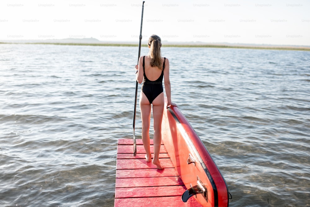 Mulher bonita de pé para trás no cais com paddleboard e remo desfrutando da vista da manhã no lago