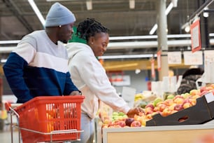 Vista lateral de una mujer joven que toma una manzana roja madura fresca y la pone en una bolsa de celofán mientras está de pie junto a su novio con el carrito de compras