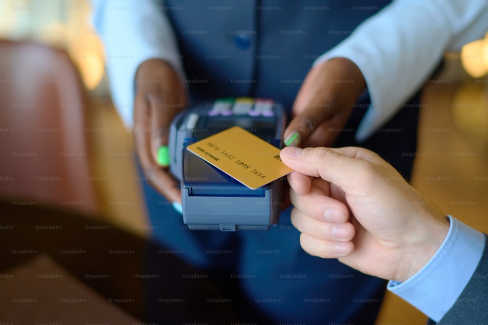 Jeune femme noire en uniforme tenant un terminal de paiement tandis qu’un homme d’affaires avec une carte de crédit payant pour un achat ou un service