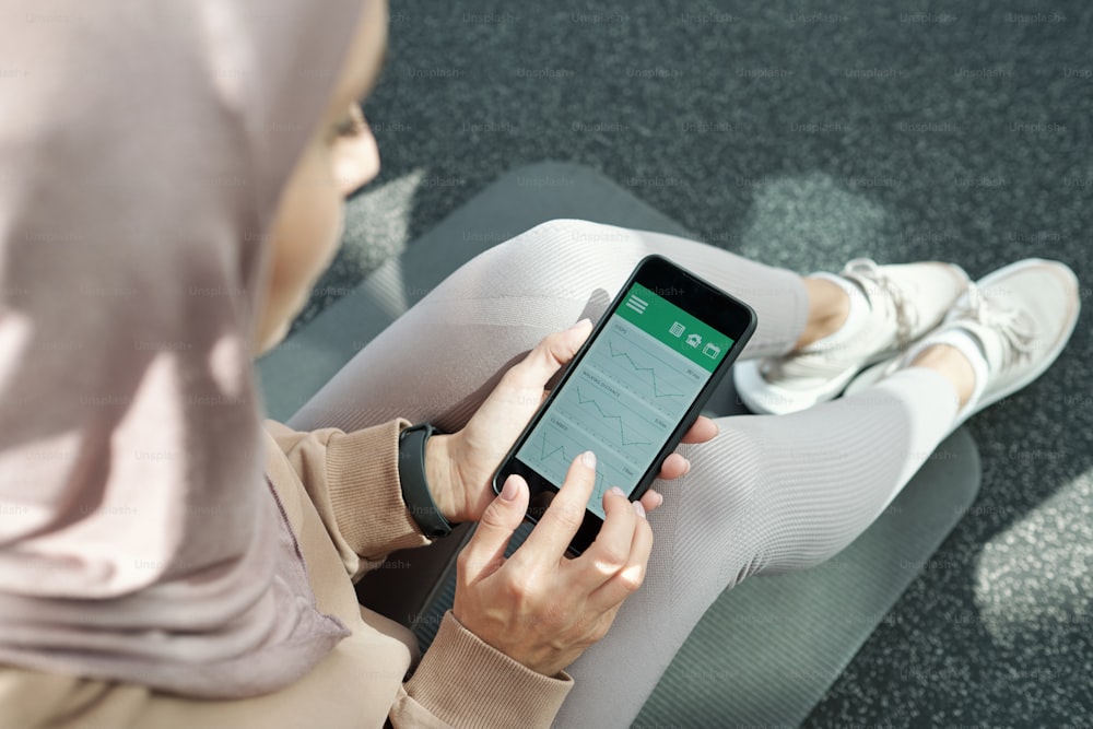 Toma de vista de ángulo alto de una mujer musulmana adulta joven moderna con pañuelo para la cabeza sentada en una colchoneta en el gimnasio mirando la pantalla del teléfono inteligente con la aplicación de fitness encendida