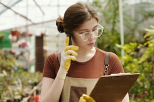 Young female florist in eyeglasses checking the availability of flowers according to document while talking to customer on phone