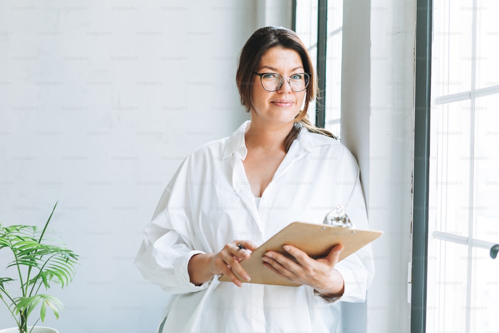 Young smiling brunette woman plus size in white shirt near window with pen and documents in hands in the bright modern office