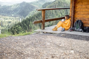 若い女性は素晴らしい山の風景を楽しみ、ハイキングのために昇華した食べ物を食べながら、木製のテラスでリラックスして座っています。旅と自然への逃避のための食べ物のコンセプト