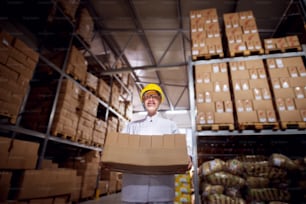 Jovem trabalhadora tensa em panos estéreis e capacete amarelo está carregando uma pilha muito pesada de caixas de papelão marrom de uma sala de armazenamento da instalação.